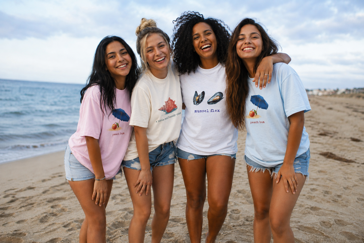 Four women standing on a beach wearing matching t-shirts with graphic designs.